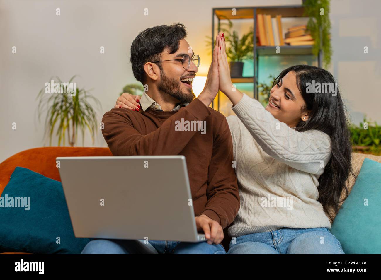 Allegra giovane e variegata coppia in abiti casual con laptop e sorpresa con la lotteria vincente per e-mail sul divano nel soggiorno di casa. La famiglia indiana felice dà cinque pugni stringenti in appartamento. Foto Stock
