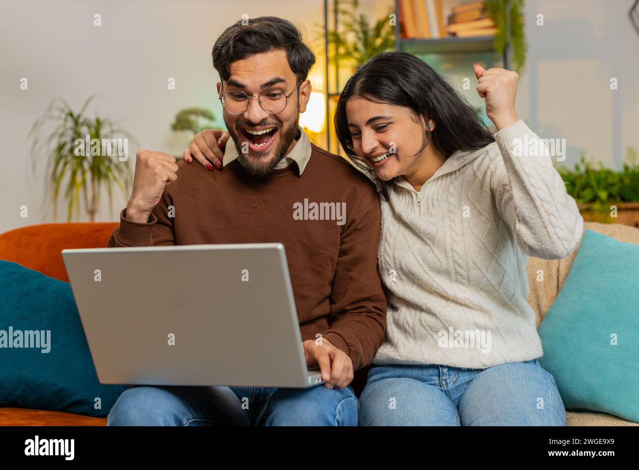Allegra giovane e variegata coppia in abiti casual che usa il computer portatile sorpreso con il jackpot di gioco online della lotteria che ha vinto per e-mail sul divano nel soggiorno di casa. Felice famiglia ispanica che stringe pugni in appartamento Foto Stock