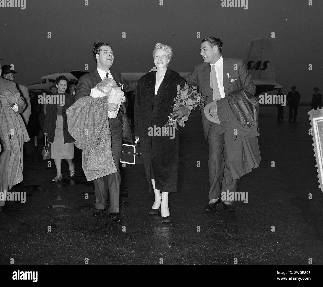 5 ottobre 1954. Amsterdam, Paesi Bassi. La star del cinema Errol Flynn (a destra) e la diciassettenne regina di bellezza danese Grethe Hoffenblad in transito a Schiphol Foto Stock