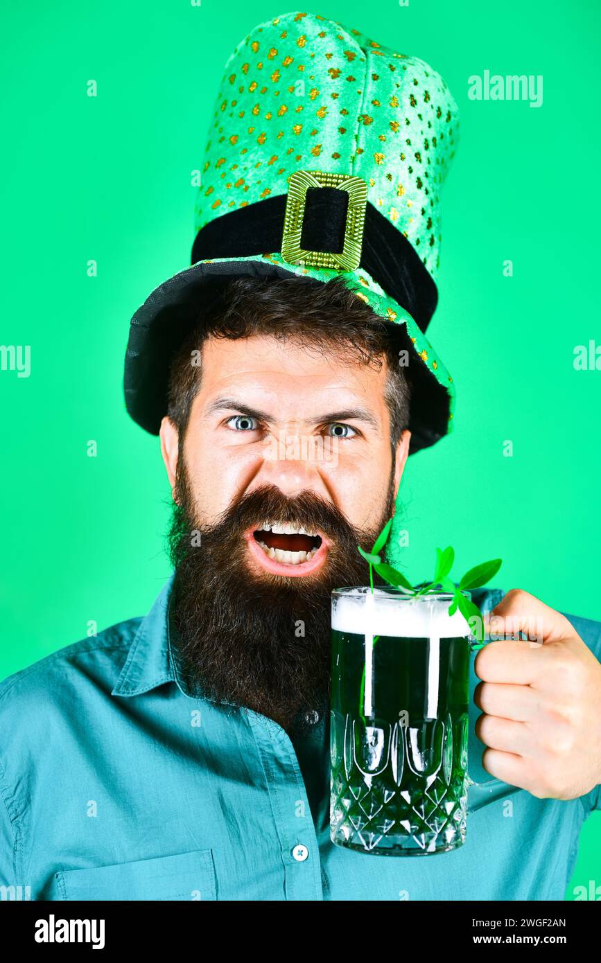Festa di San Patrizio. L'uomo barbuto in cappello da leprechaun tiene birra verde di vetro con trifoglio. Primo piano ritratto di uomo arrabbiato con vestiti verdi Foto Stock