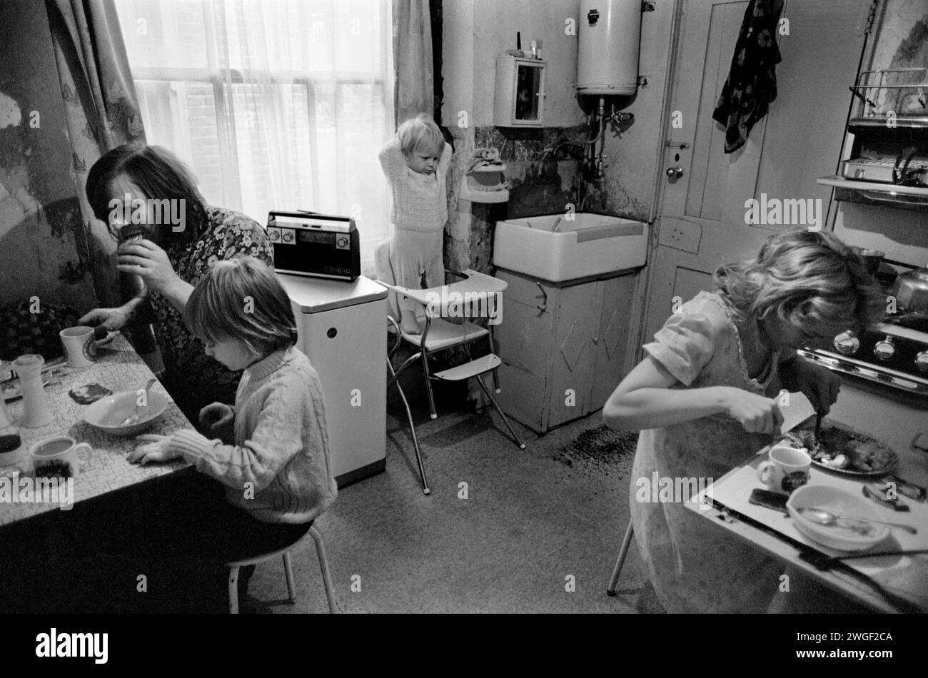 Famiglia della classe operaia 1970s Regno Unito, a casa Fulham Londra 1972. Questa è la loro cucina. Non avevano un bagno privato che era condiviso con altre famiglie della casa. Il lavandino del maggiordomo viene utilizzato per il lavaggio del viso al mattino. Il tavolo della cucina non è abbastanza grande da permettere a tutti di sedersi. Mamma usa il piccolo "tavolo" a scomparsa quando mangiano tutti insieme. Bambino in seggiolone. HOMER SYKES Foto Stock