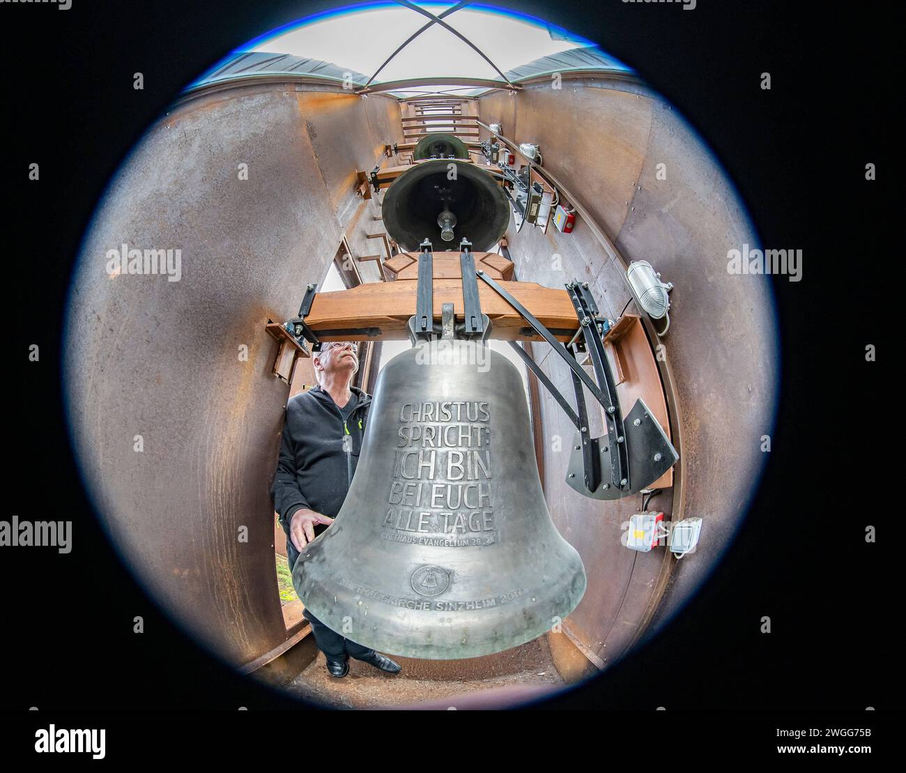 Auf den Kopf gestellter Glockenturm der evangelischen Matthaeusgemeinde in Sinzheim foto vom 25.01.2024: Kirchenaeltester Helmut Huber neben der untersten und groessten Bronzeglocke mit der Inschrift Christus spricht: ICH bin bei euch alle Tage . Kirchtuerme sind meist von weitem sichtbar und mit ihrem Glockengelaeut hoerbar. SIE zeigen den Menschen, wo sie sich zu Gebet und Gottesdienst treffen koennen. Ein ungewoehnliches Projekt aus Karlsruhe stellt Glockentuerme einfach auf den Kopf. Era merkwuerdig erscheint, Hat einen praktischen Grund: In Zeiten knapper Kassen koennen so baufaellige ode Foto Stock