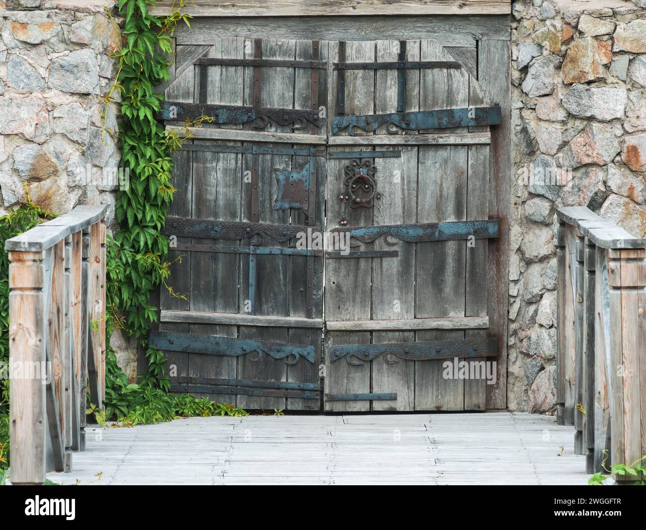 Vecchio cancello in legno con cerniere forgiate in un muro di pietra ricoperto di edera e un ponte con ringhiere in legno davanti ad esso Foto Stock