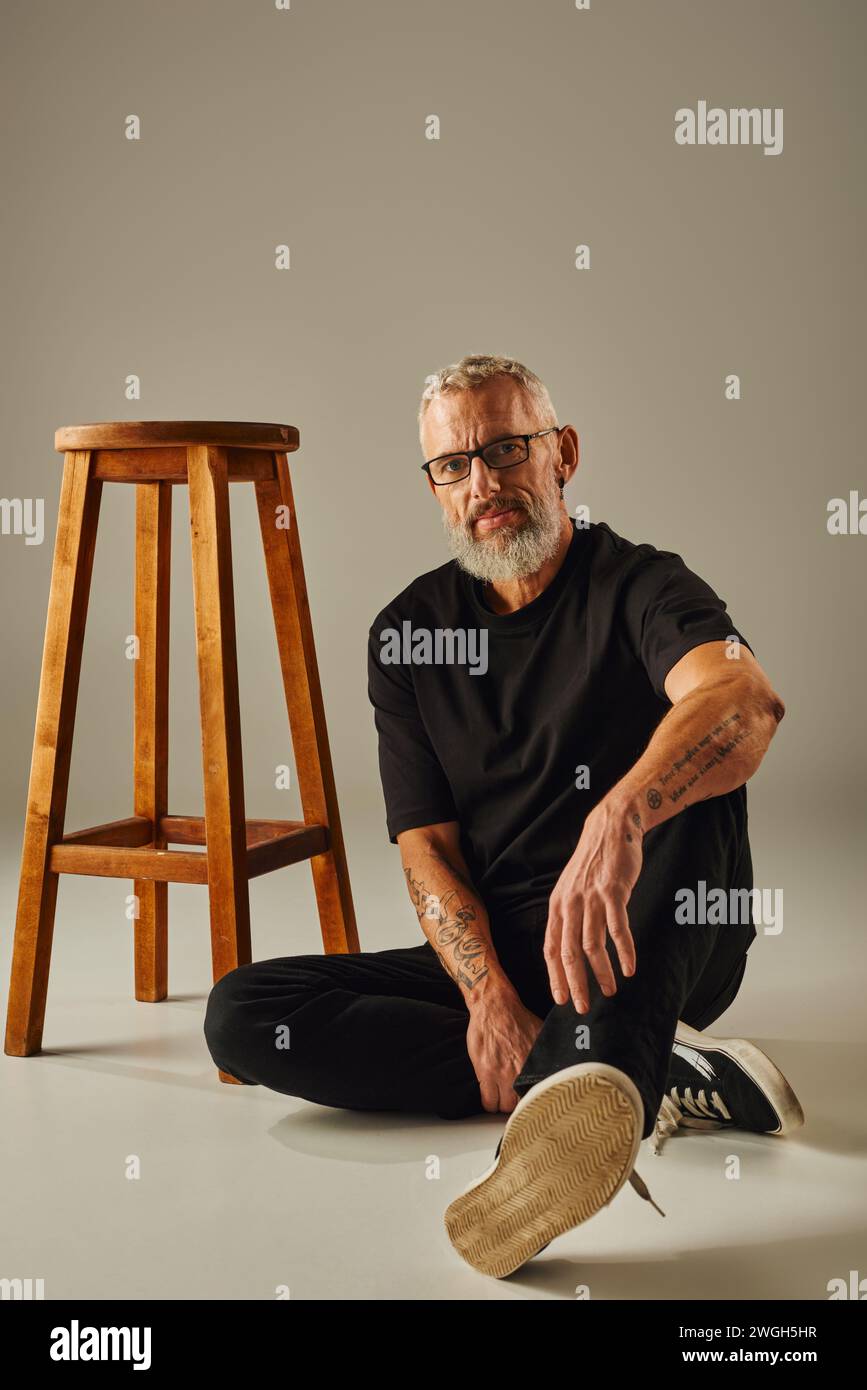 attraente uomo barbuto maturo in t shirt nera con tatuaggi seduti sul pavimento e che guarda la macchina fotografica Foto Stock