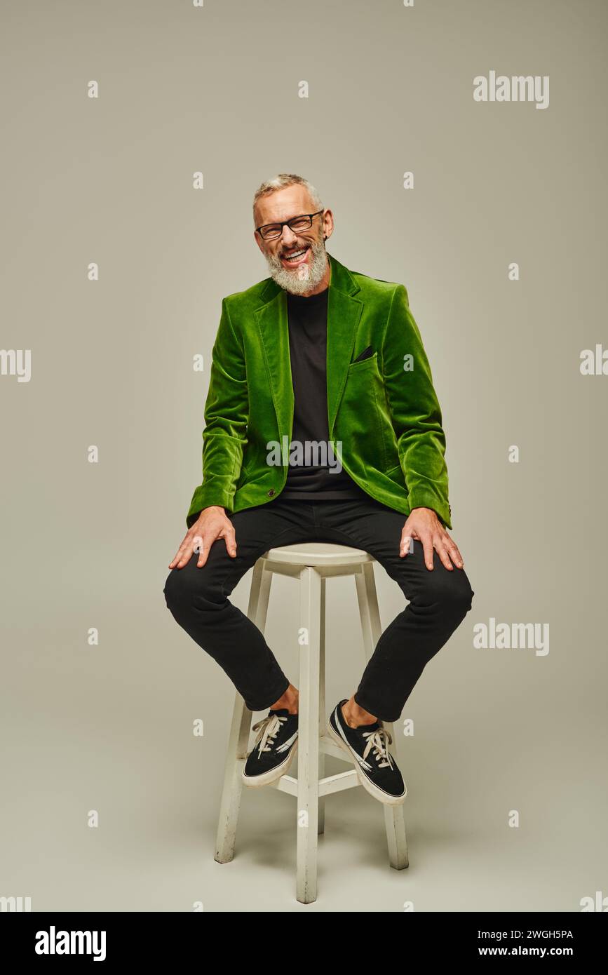 allegro barbuto attraente uomo maturo in blazer verde seduto su una sedia alta e sorridente alla fotocamera Foto Stock