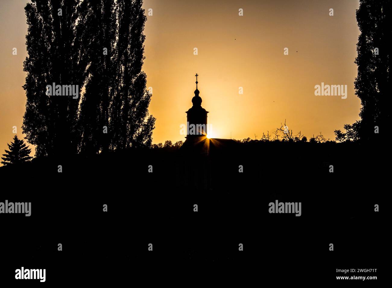 Silhouette drammatica di una chiesa con una croce al tramonto a Baranya, Ungheria, il sole che sbuca da dietro l'edificio, incorniciato da alberi scuri Foto Stock