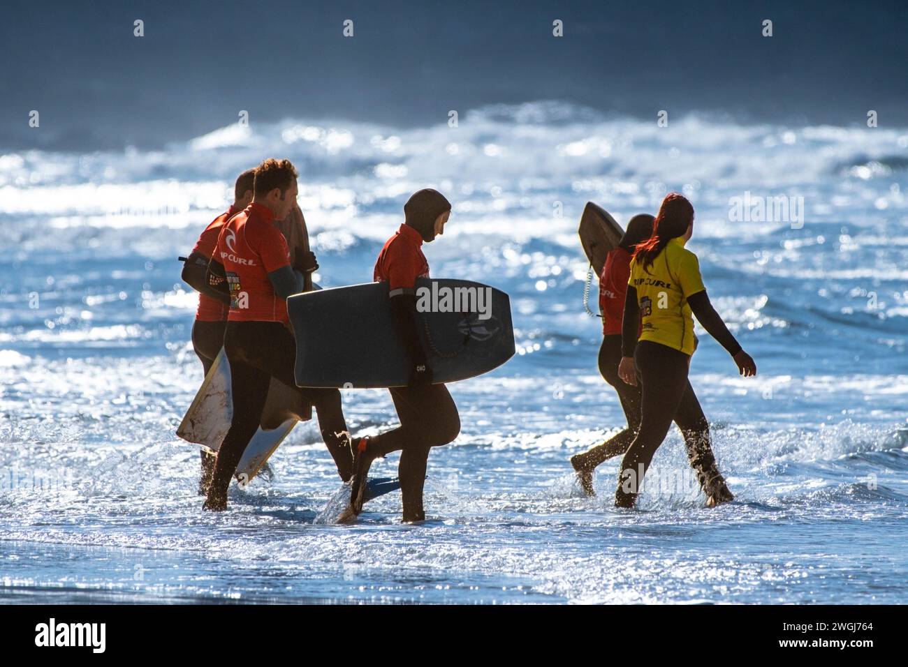 I visitatori e un istruttore di surf camminano in mare per una lezione di body boarding a Fistral Beach a Newquay in Cornovaglia nel Regno Unito. Foto Stock