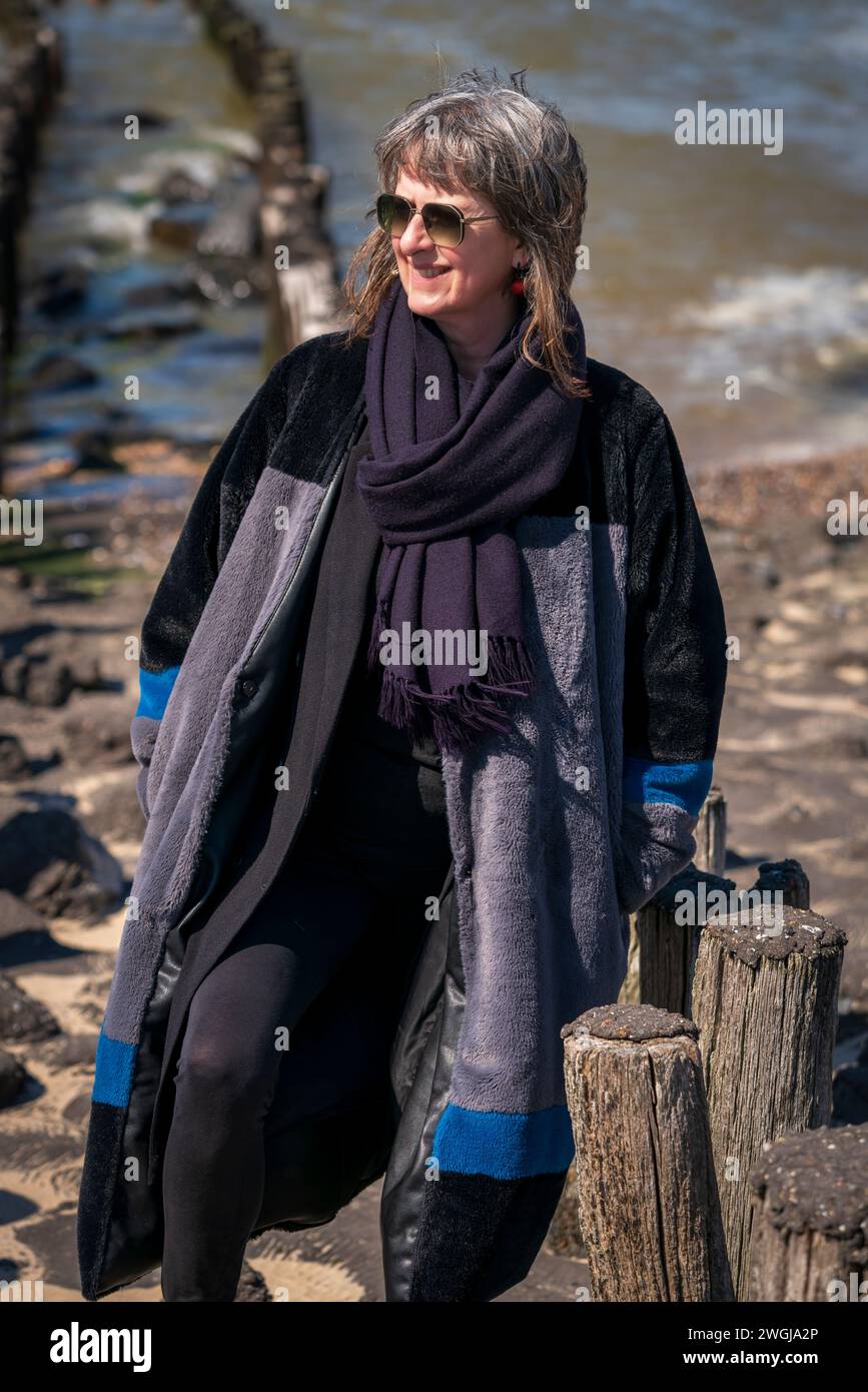 Una donna matura vestita con abiti invernali sorride mentre passeggia sulla costa olandese con il mare alle spalle che affronta la giornata fredda. Foto Stock