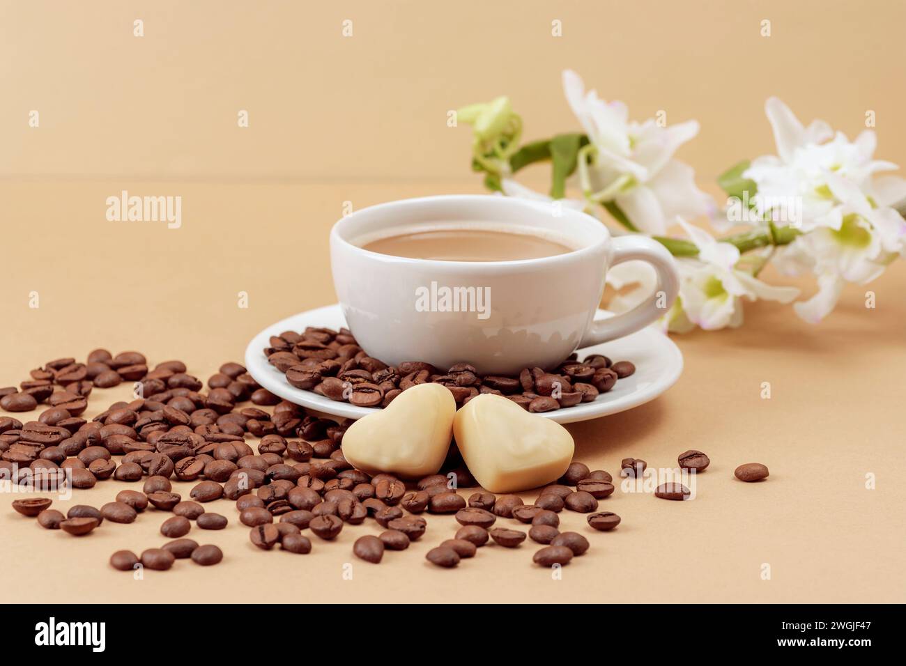 Tazza di caffè e chicchi di caffè, caramelle a forma di cuore e fiori bianchi su sfondo beige. Il giorno di San Valentino. Primo piano. Foto Stock