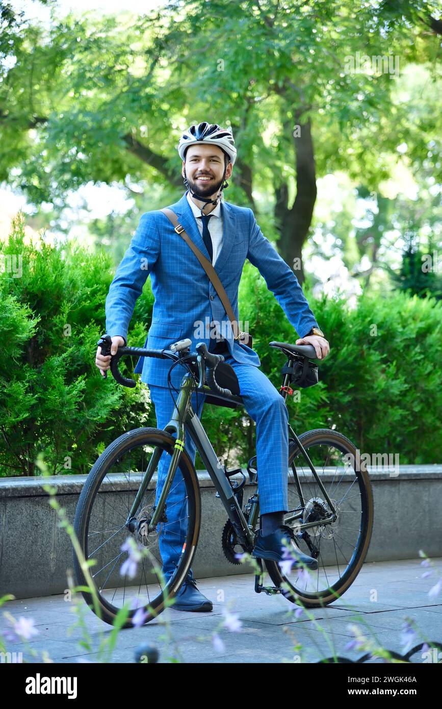 Uomo d'affari con barba in tuta e casco con bici in piedi nel parco. Concetto di business e stile urbano. Foto Stock