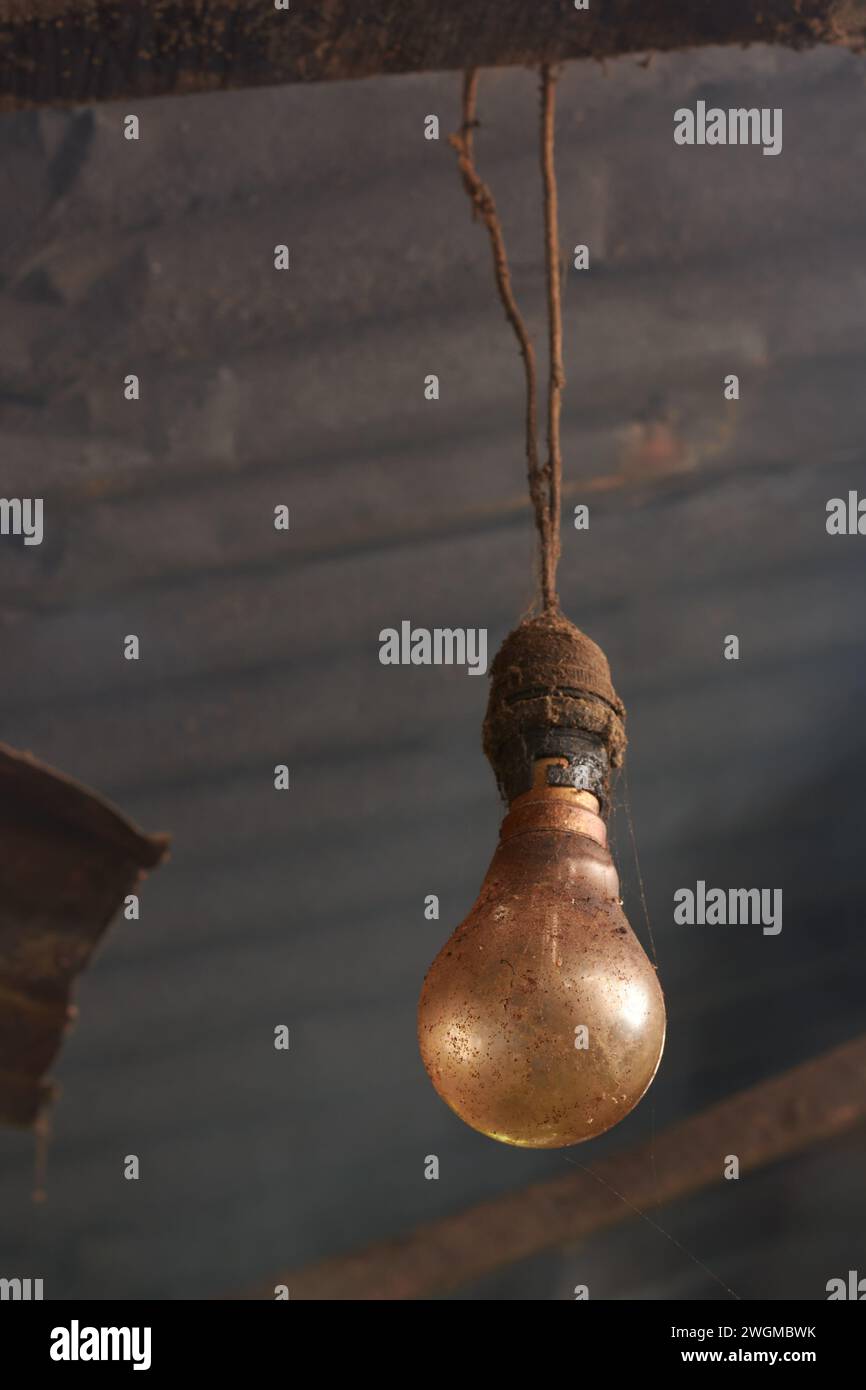 primo piano di una vecchia lampadina a incandescenza sporca o polverosa, appesa al cavo elettrico con tetto in metallo e fumo sullo sfondo, lampada da cucina del villaggio Foto Stock