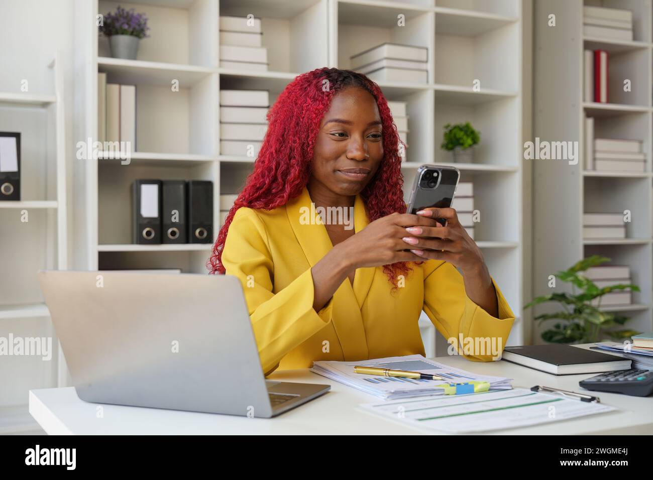 Donna d'affari africana che utilizza l'app per smartphone per giocare, fare shopping online e ordinare la consegna mentre lavora su un computer portatile nel Foto Stock