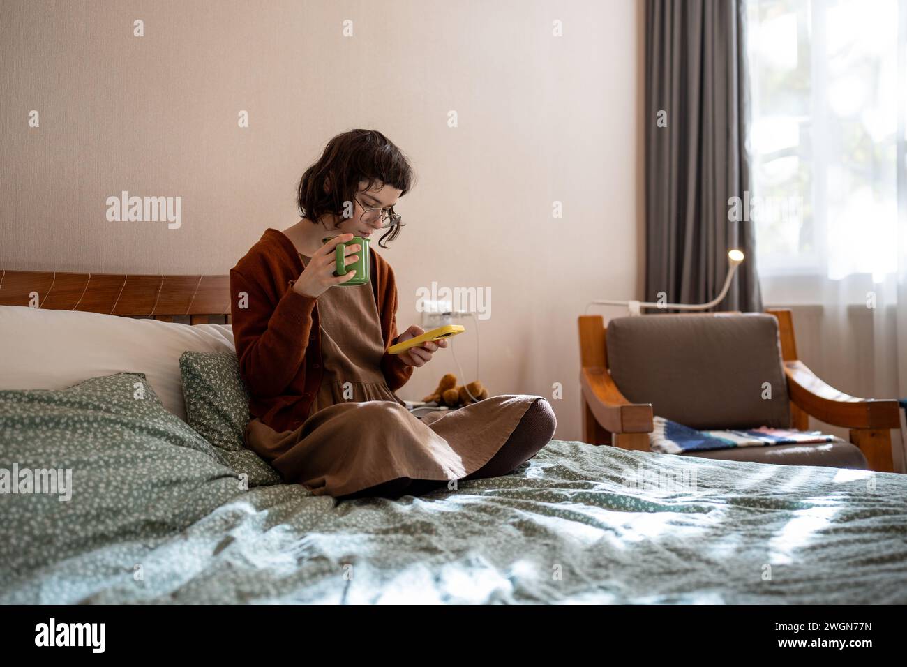 Donna pensiva seduta sul letto con una tazza di tè, leggendo notizie su Internet, appesa ai social network Foto Stock