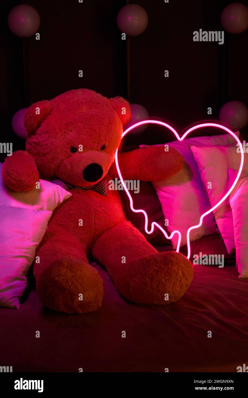 Un simpatico orsacchiotto marrone giace nei cuscini, contiene un cuore rosa neon luminoso. San Valentino 14 febbraio, Gift Romantic background. Dichiarazione di Foto Stock