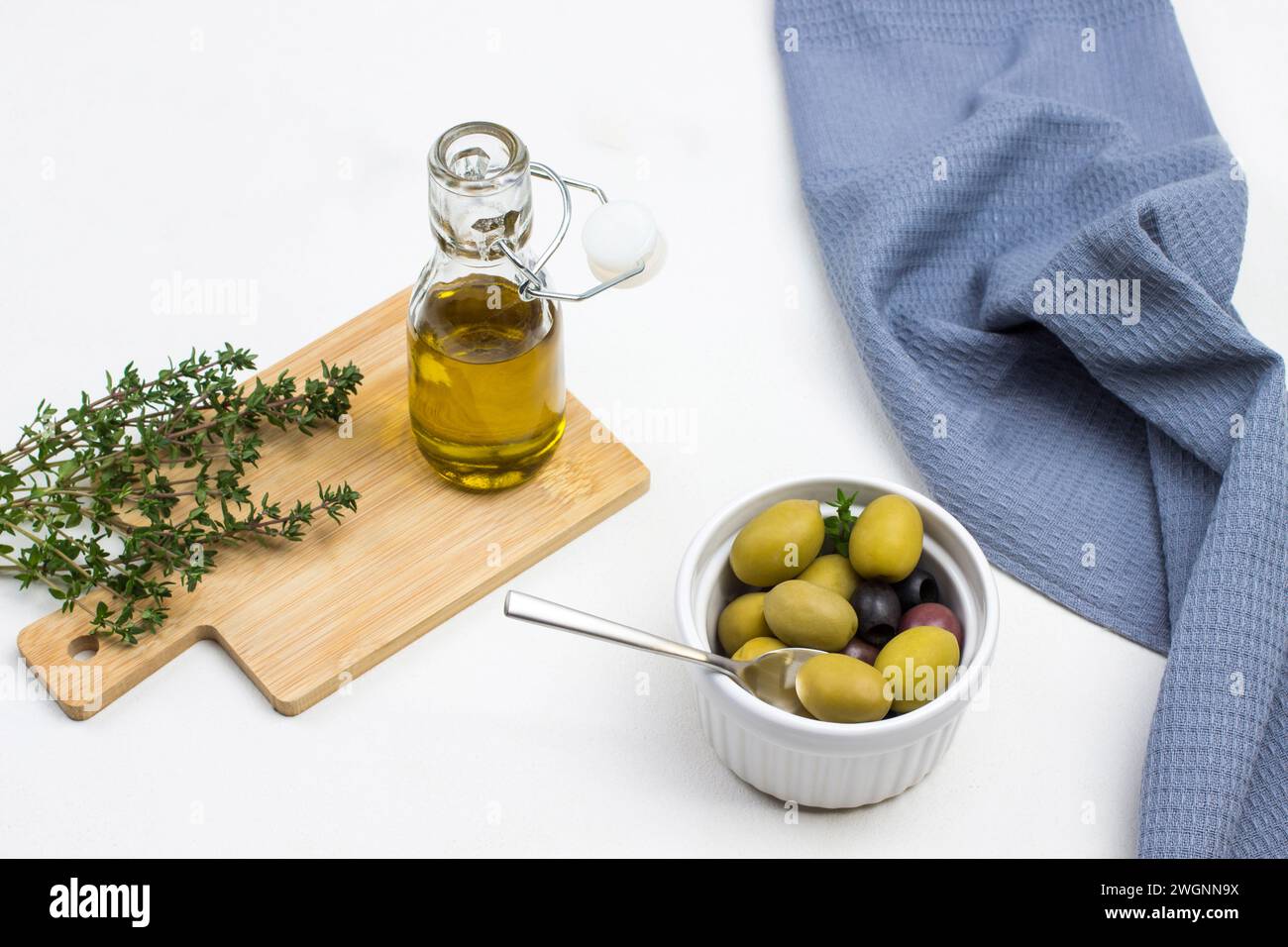 Bottiglia di olio d'oliva e ciuffi di timo a bordo. Olive in un recipiente di ceramica. Tovagliolo grigio sul tavolo. Sfondo bianco. Vista dall'alto. Foto Stock
