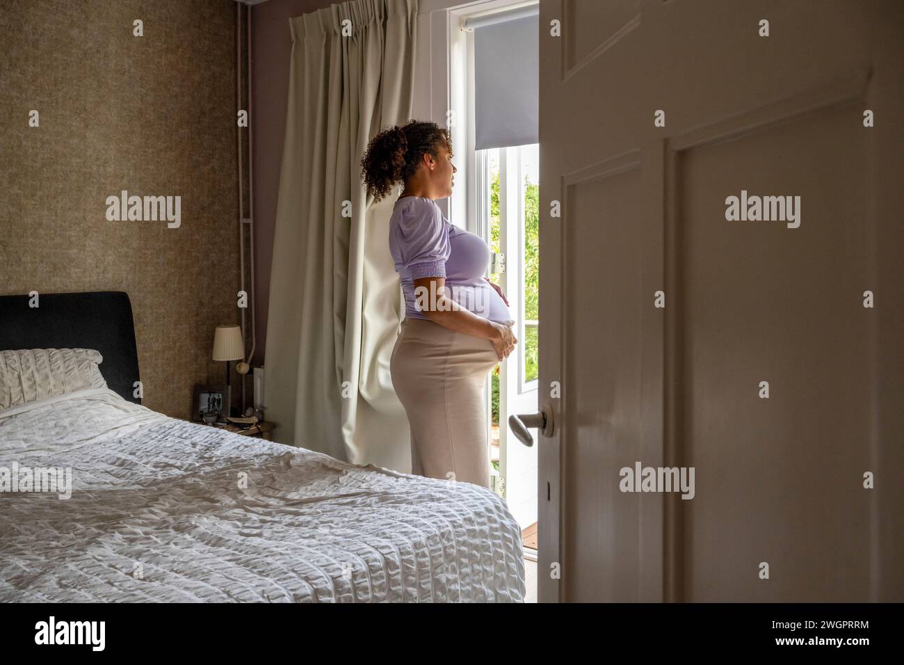 Donna incinta afroamericana che si rilassa guardando fuori dalla finestra della sua camera da letto Foto Stock