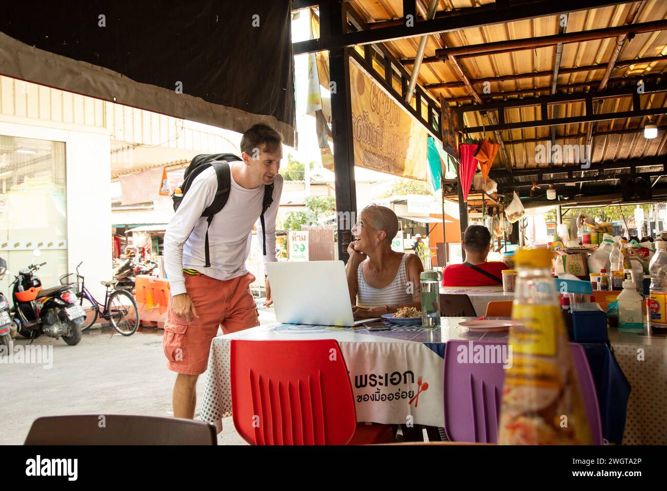 Una donna di mezza età che viaggia da sola, che lavora con il suo laptop in un ristorante locale, si ferma per chiacchierare con un altro viaggiatore maschile. Foto Stock
