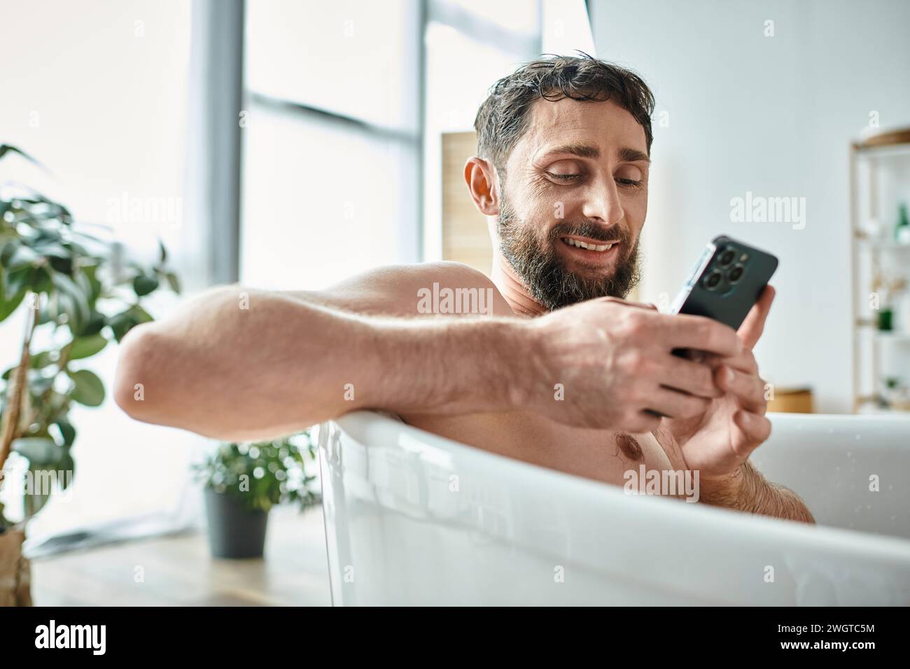 uomo bello e allegro con barba che guarda il suo smartphone mentre è in vasca da bagno, consapevolezza della salute mentale Foto Stock