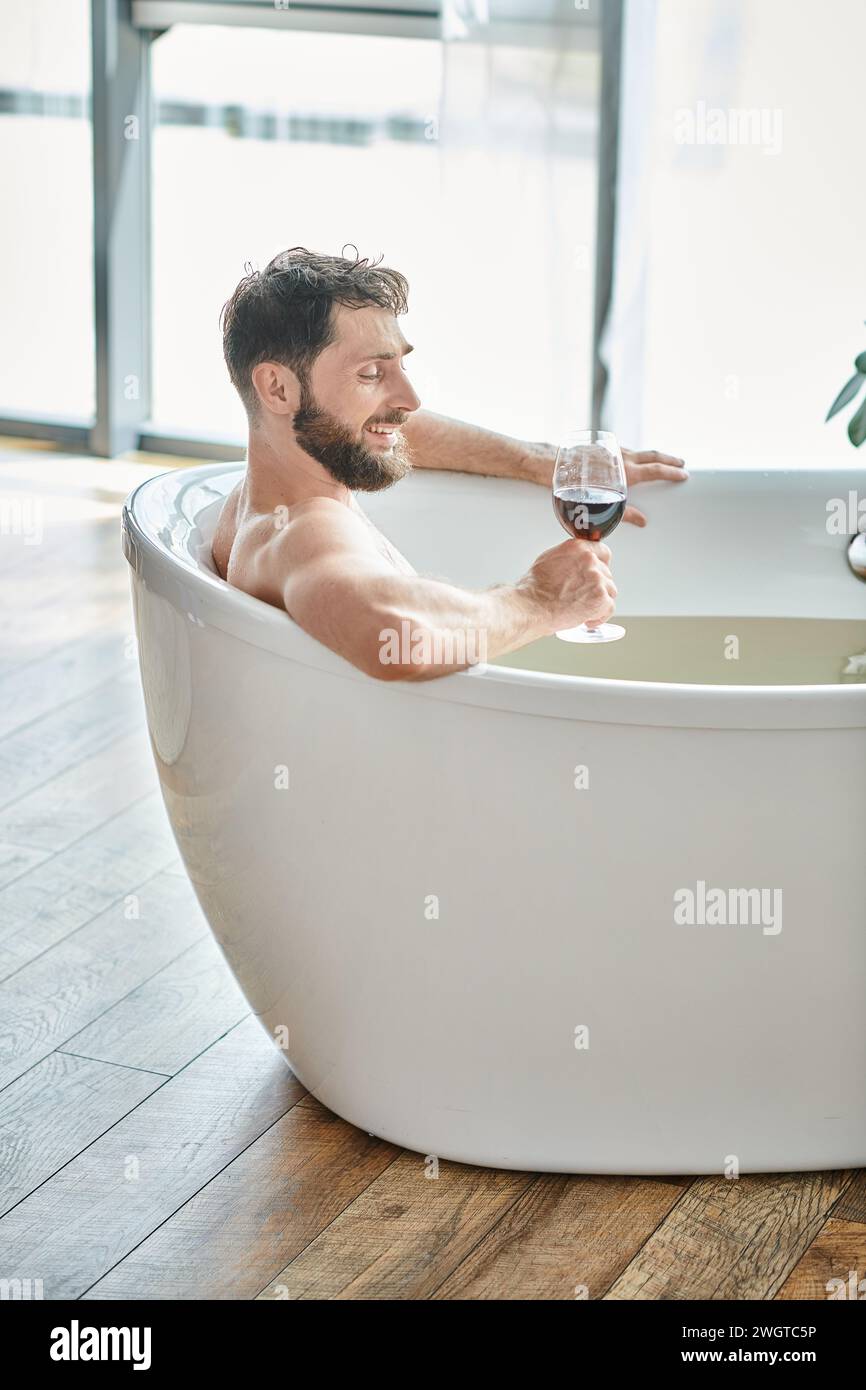 uomo gioioso e bello con barba che si rilassa nella vasca da bagno con un bicchiere di vino rosso, consapevolezza della salute mentale Foto Stock