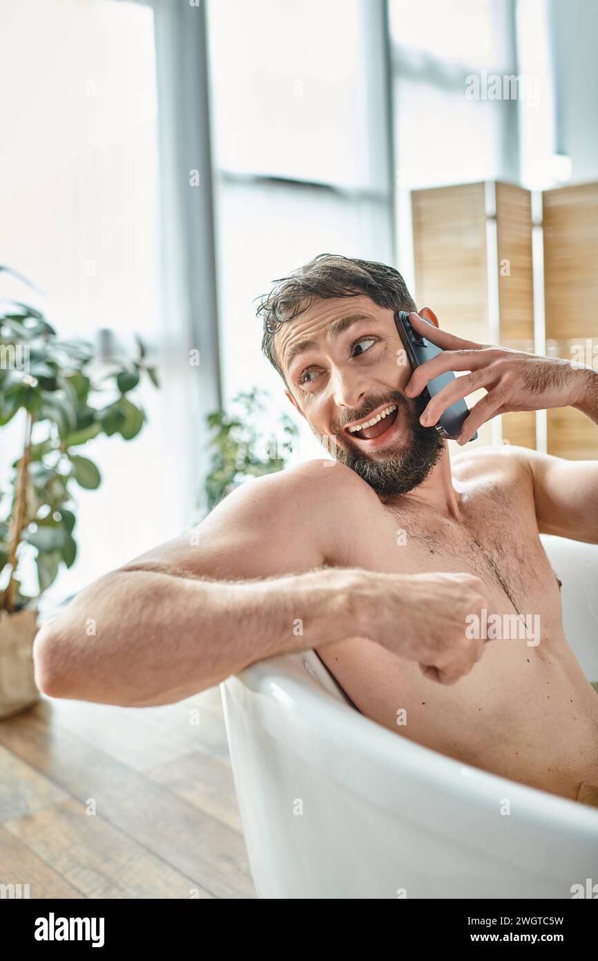 uomo bello e gioioso con barba che parla al telefono mentre si rilassa nella vasca da bagno, salute mentale Foto Stock