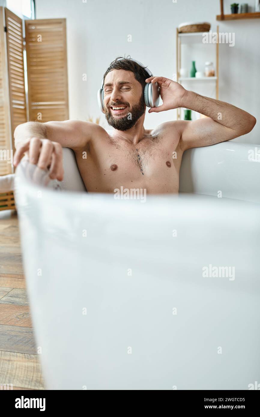 uomo gioioso e attraente con barba e cuffie seduti e rilassanti nella vasca da bagno, salute mentale Foto Stock