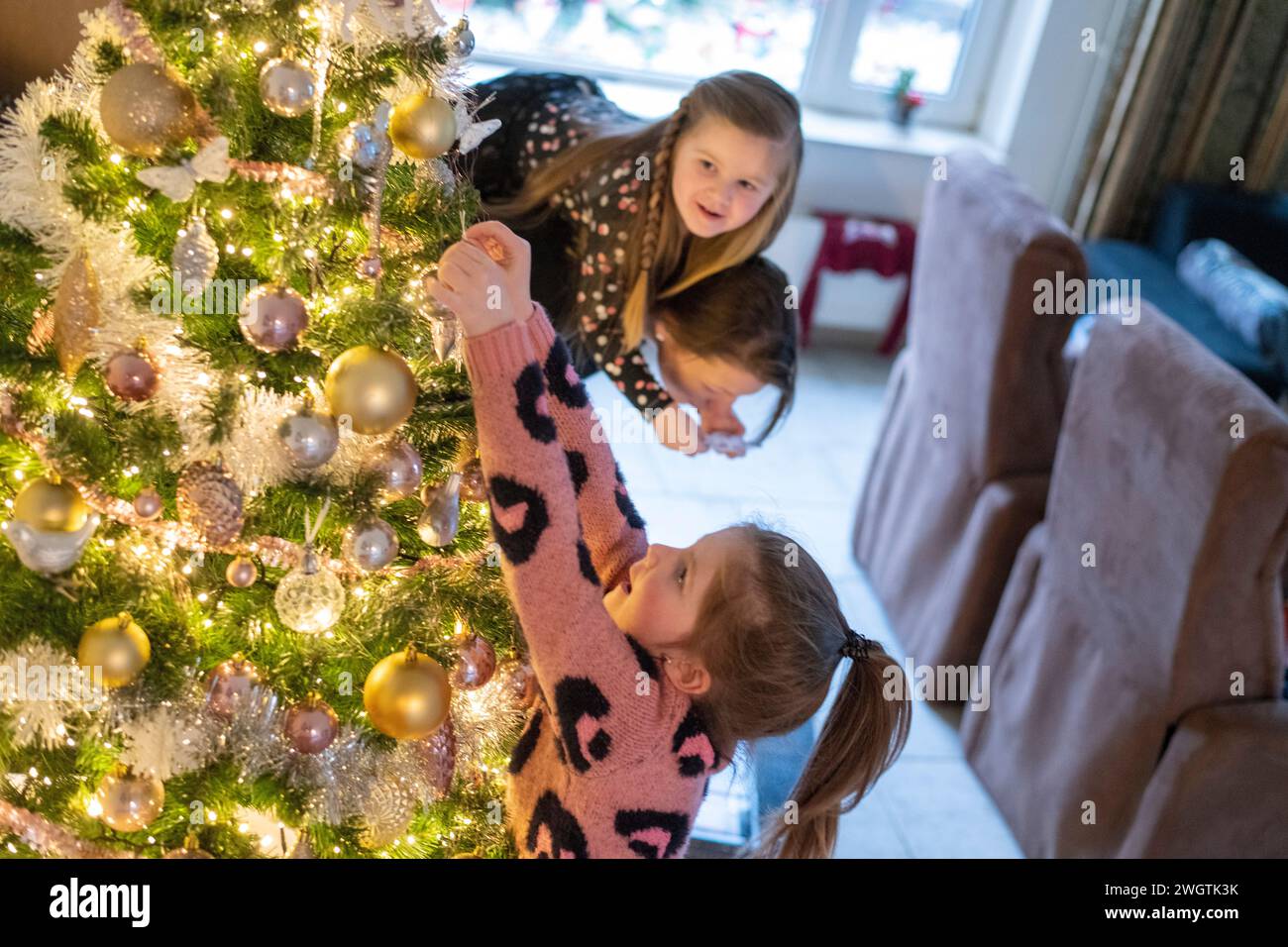Bambina che sale sulla cima delle spalle della madre per mettere gli ornamenti sul loro albero di Natale. Sua sorella si sta avvicinando per mettere un baule Foto Stock