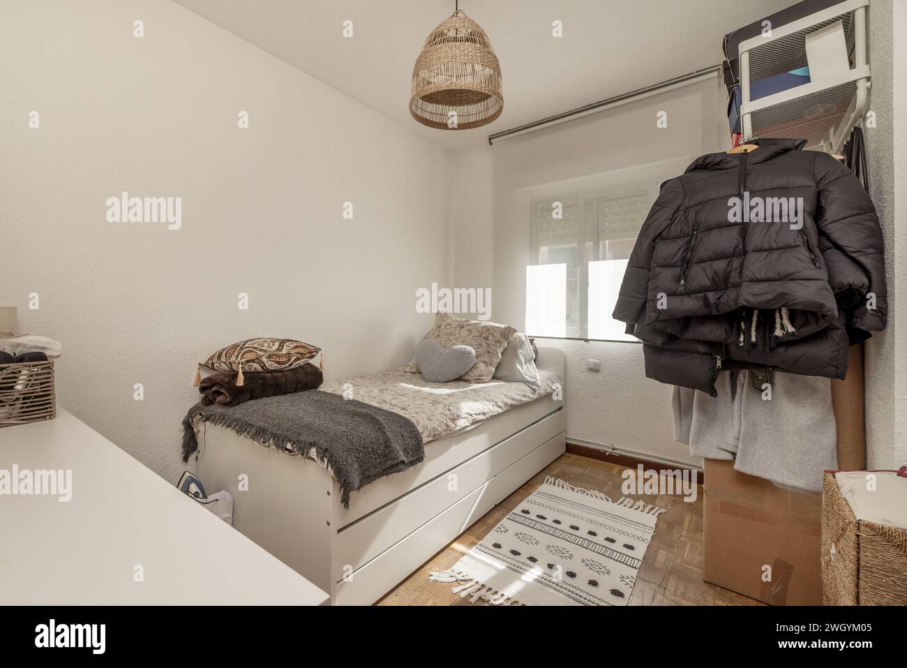 Una camera da letto per giovani con un letto singolo, radiatore in alluminio, abiti appesi su un ripiano, una finestra con una tendina avvolgibile e una cassettiera bianca con molti cassetti Foto Stock