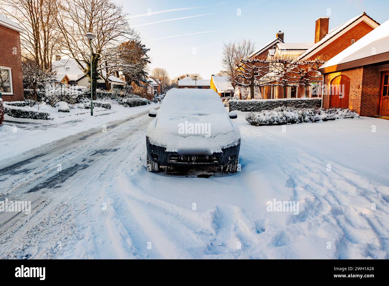 Paesaggio urbano invernale con una strada urbana, marciapiedi e un'auto coperta da uno spesso strato di neve, cespugli ghiacciati e alberi nudi su sfondo sfocato, soleggiato Foto Stock