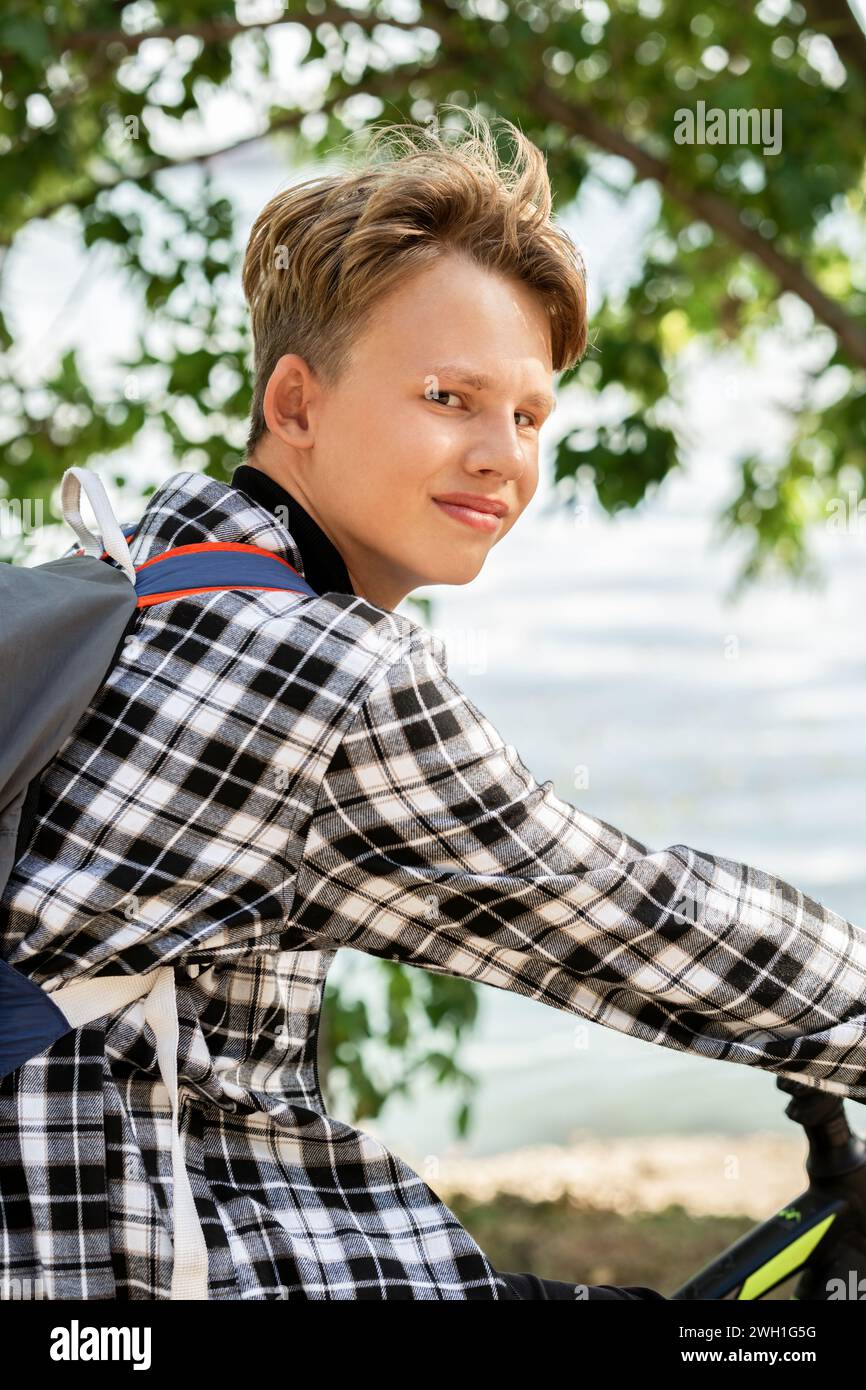 Ritratto di un ragazzo sorridente con i capelli biondi in camicia a quadri con zaino seduto in bicicletta, su alberi sfocati e sullo sfondo riflettente del laghetto nelle giornate di sole Foto Stock