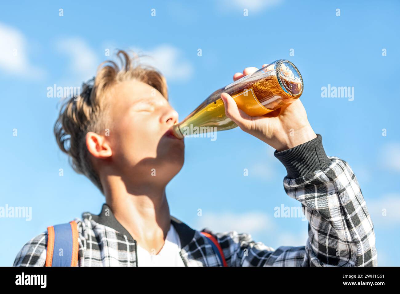 Un ragazzo gioioso beve limonata da una bottiglia di vetro sullo sfondo di un bellissimo cielo. Indossa una camicia a quadri e uno zaino. Colori vivaci Foto Stock