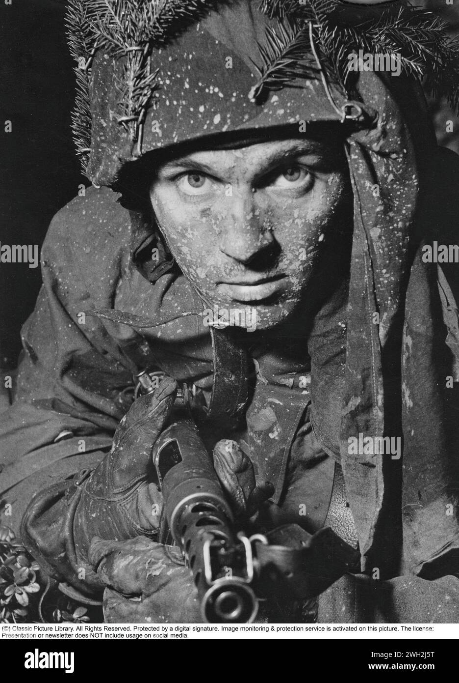 Soldato svedese negli anni '1950 Un giovane uomo durante un esercizio militare si è mimetizzato bene con rami di abete nel casco. Ha in mano una mitragliatrice carica ed è pronto per la battaglia. Nel 1945, la Svezia introdusse il 9 mm Parabellum Carl Gustaf m/45 con un progetto che prese in prestito e migliorò molti elementi di progettazione dei precedenti progetti di mitragliatrici. È dotato di un serbatoio tubolare in acciaio stampato con un supporto ripiegabile laterale. L'm/45 fu ampiamente esportato, e particolarmente popolare tra gli agenti della CIA e le forze speciali statunitensi durante la guerra del Vietnam. In servizio negli Stati Uniti era conosciuto come "Swedish-K" Foto Stock