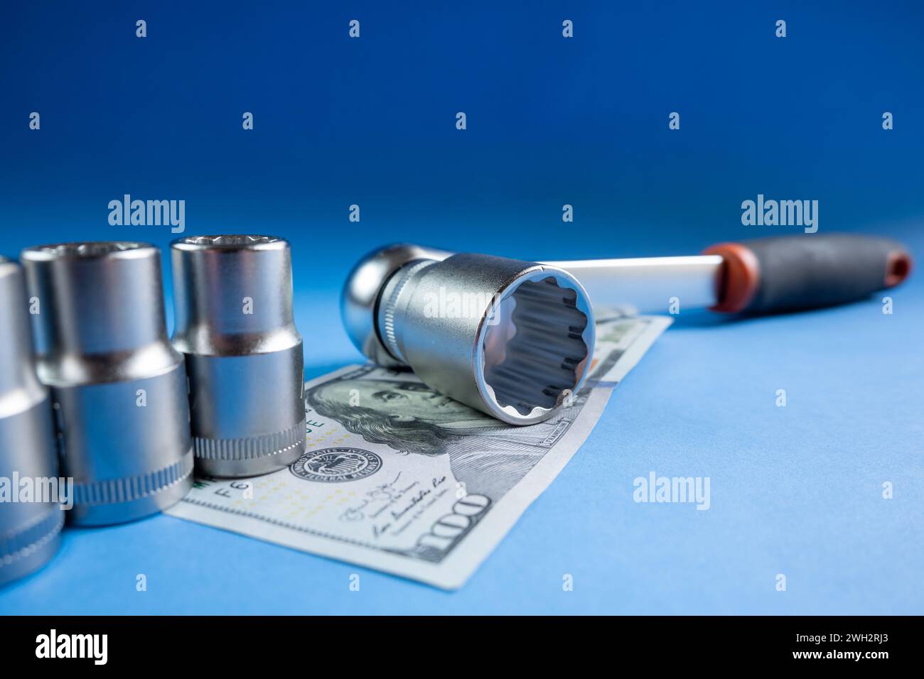 Il concetto di costo meccanico realizzato con una chiave a cricchetto su una banconota da cento dollari su sfondo blu. Foto Stock