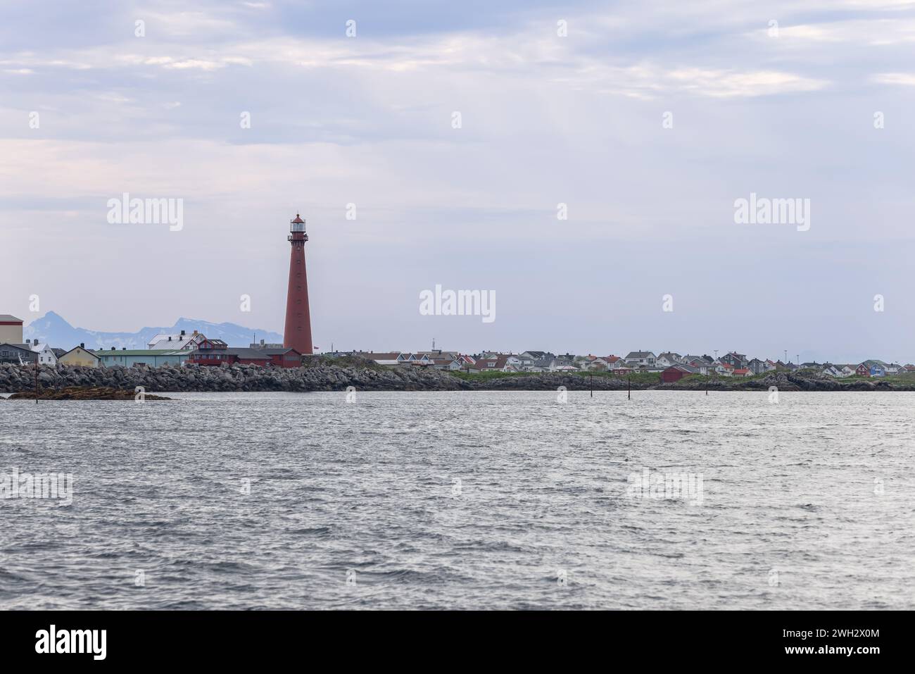 Un torreggiante faro rosso, un faro di Andenes, Norvegia, sorge sopra un mare calmo con lo sfondo di una cittadina accogliente e colorata. Foto Stock