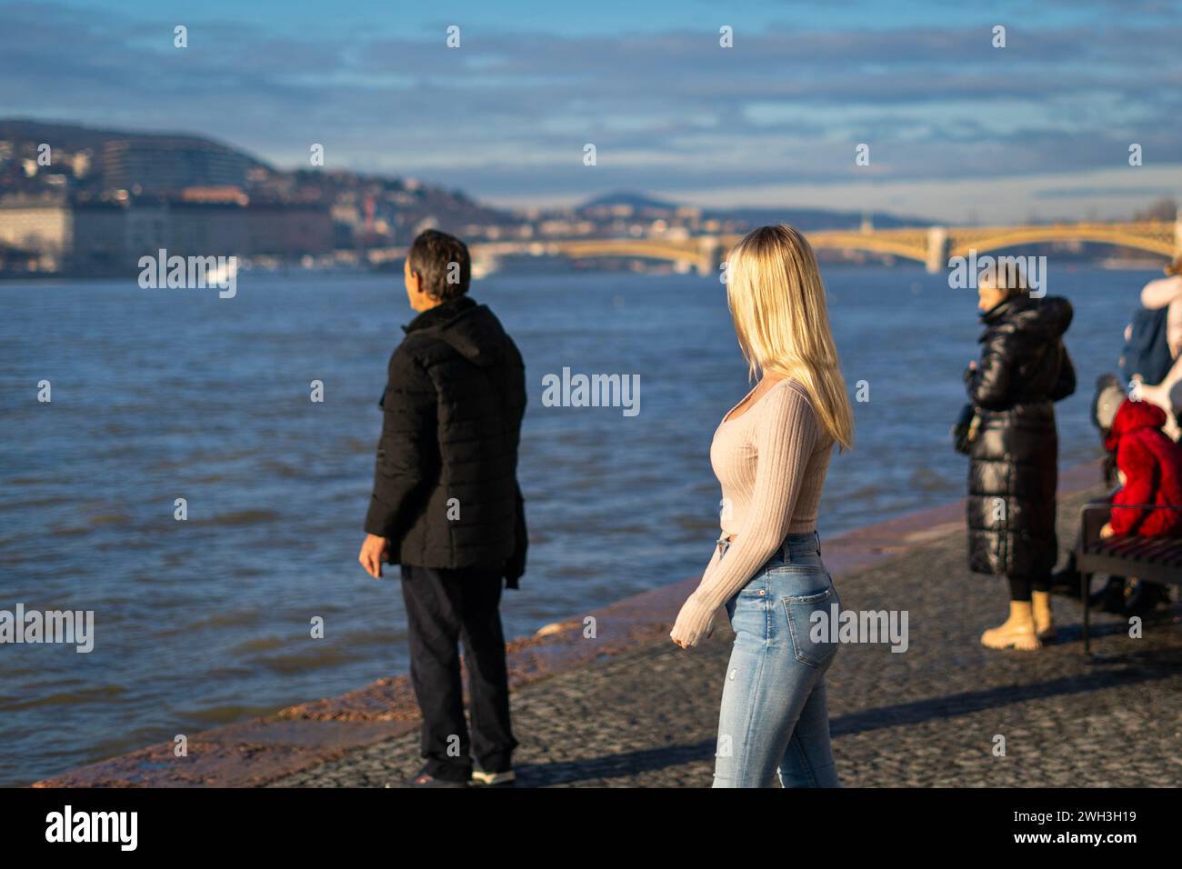 I turisti si crogiolano al sole invernale vicino al fiume Danubio allagato sul molo di Budapest Foto Stock