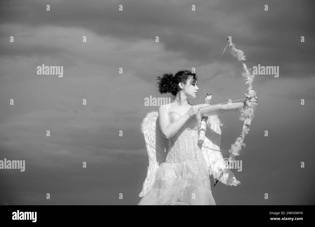 Ragazza in abito bianco angeli, vestito con ali puntate con arco e freccia su sfondo cielo. Concetto di giorno di San Valentino. Angelo teen in cielo. Foto Stock