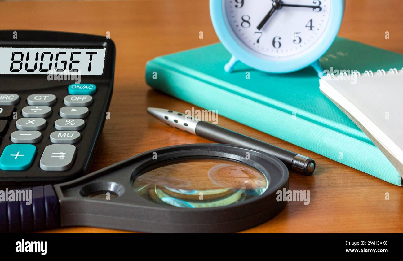 Calcolatrice con la parola BUDGET sul display. Penna, lente d'ingrandimento, orologio e blocco note su sfondo in legno. Foto concettuale Foto Stock