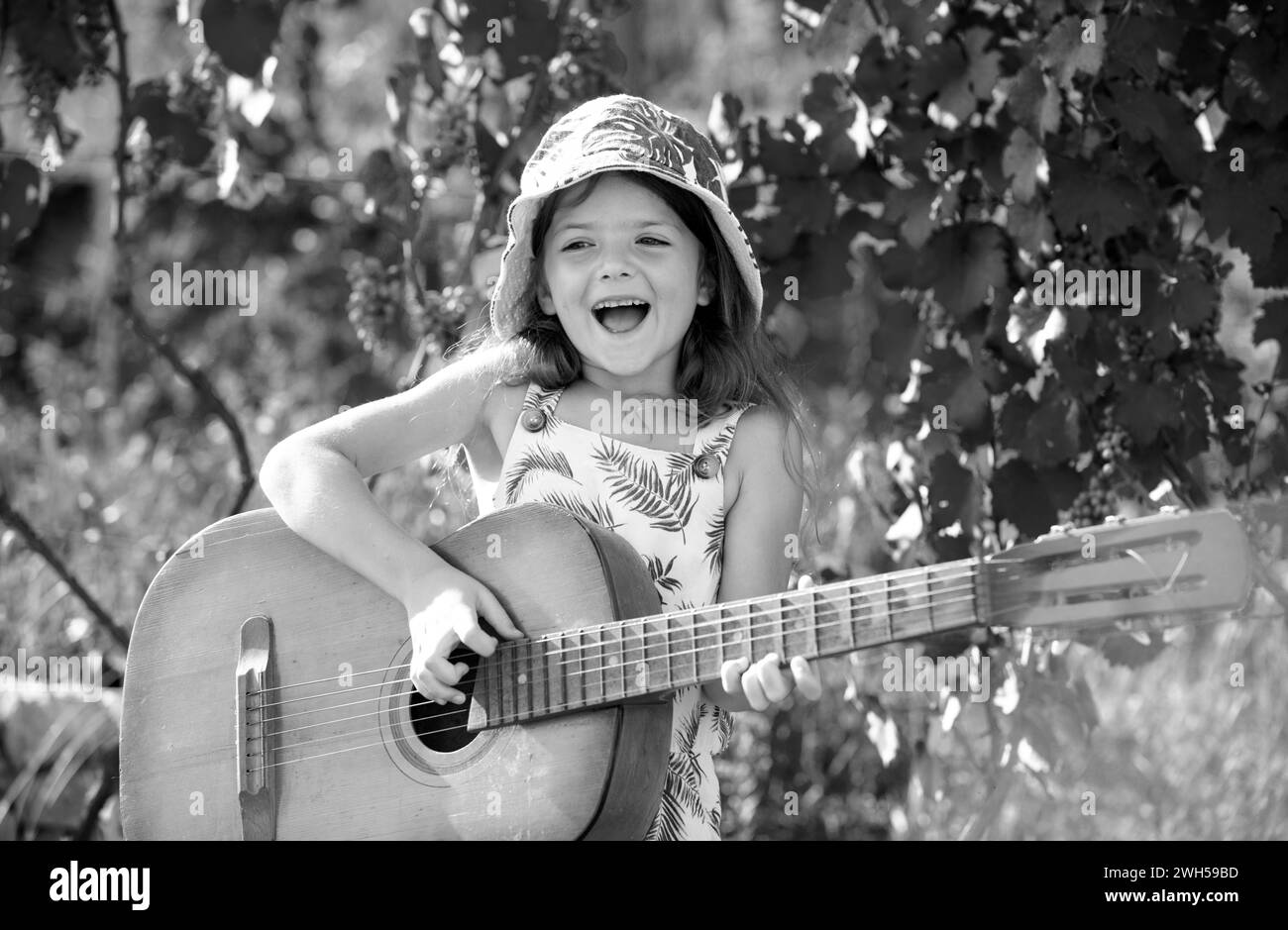 Bambino musicista che suona la chitarra. Bambina alla moda che indossa un abito estivo che si diverte sul cortile. Faccia sognante dei bambini. Foto Stock