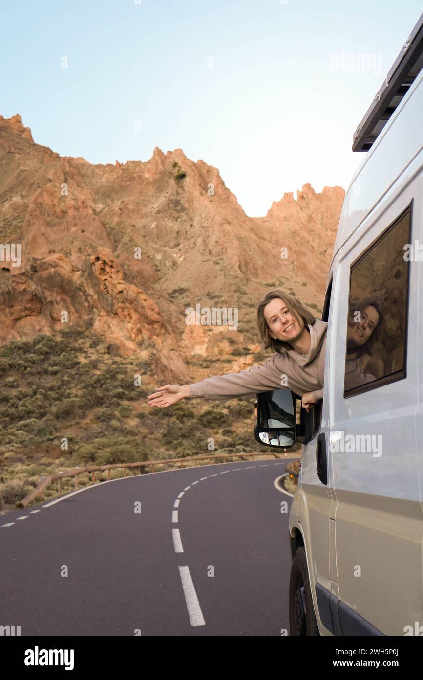 Un uomo si allunga con gioia il braccio da un furgone bianco, mentre viaggia su una tortuosa strada di montagna durante il caldo bagliore del tramonto. Foto Stock