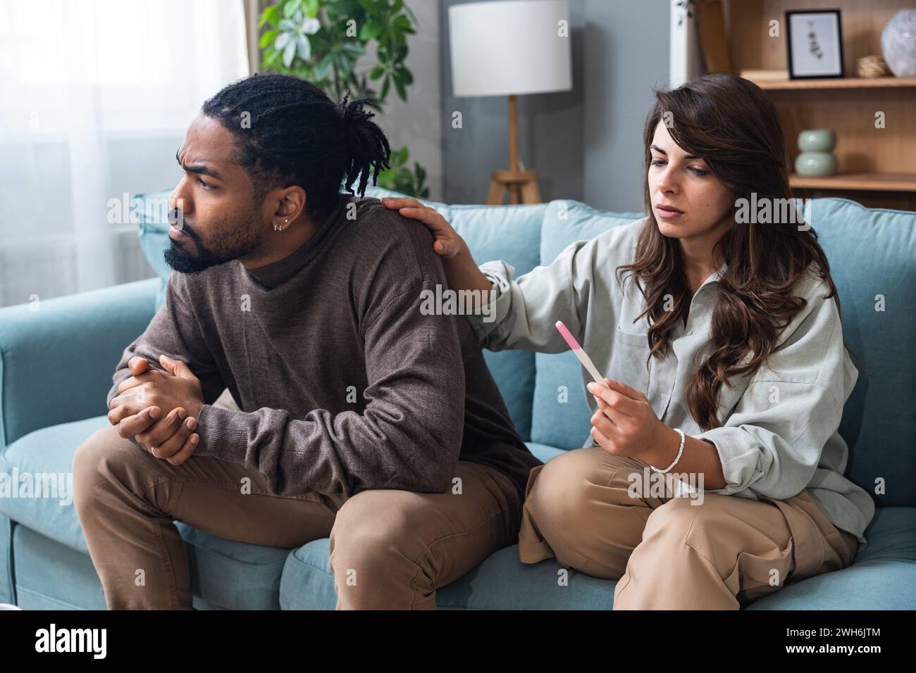 Infelice giovane donna seduta sul divano con il test di gravidanza, marito afroamericano che le urla, in casa. La famiglia Millennial ha un conflitto per un Foto Stock