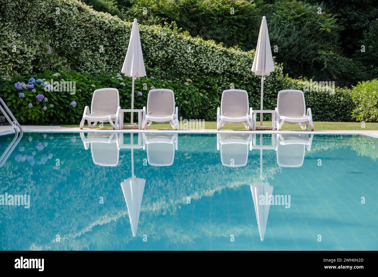 Lettini e ombrelloni bianchi vuoti che si riflettono in una piscina blu Foto Stock