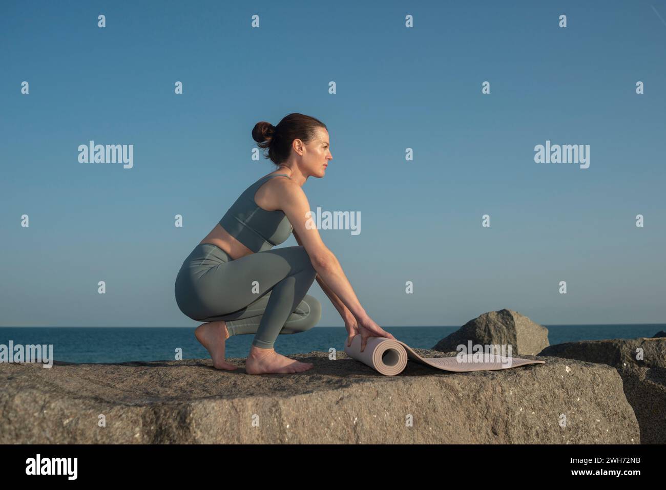 donna sportiva che srotola un tappetino da ginnastica su una roccia vicino all'oceano Foto Stock