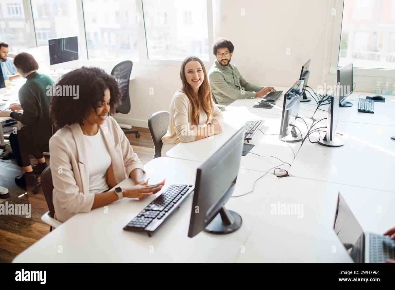 Un team di professionisti lavora in un ufficio open space luminoso. Il gruppo diversificato si concentra sugli schermi dei computer, suggerendo un ambiente di lavoro collaborativo e esperto di tecnologia Foto Stock