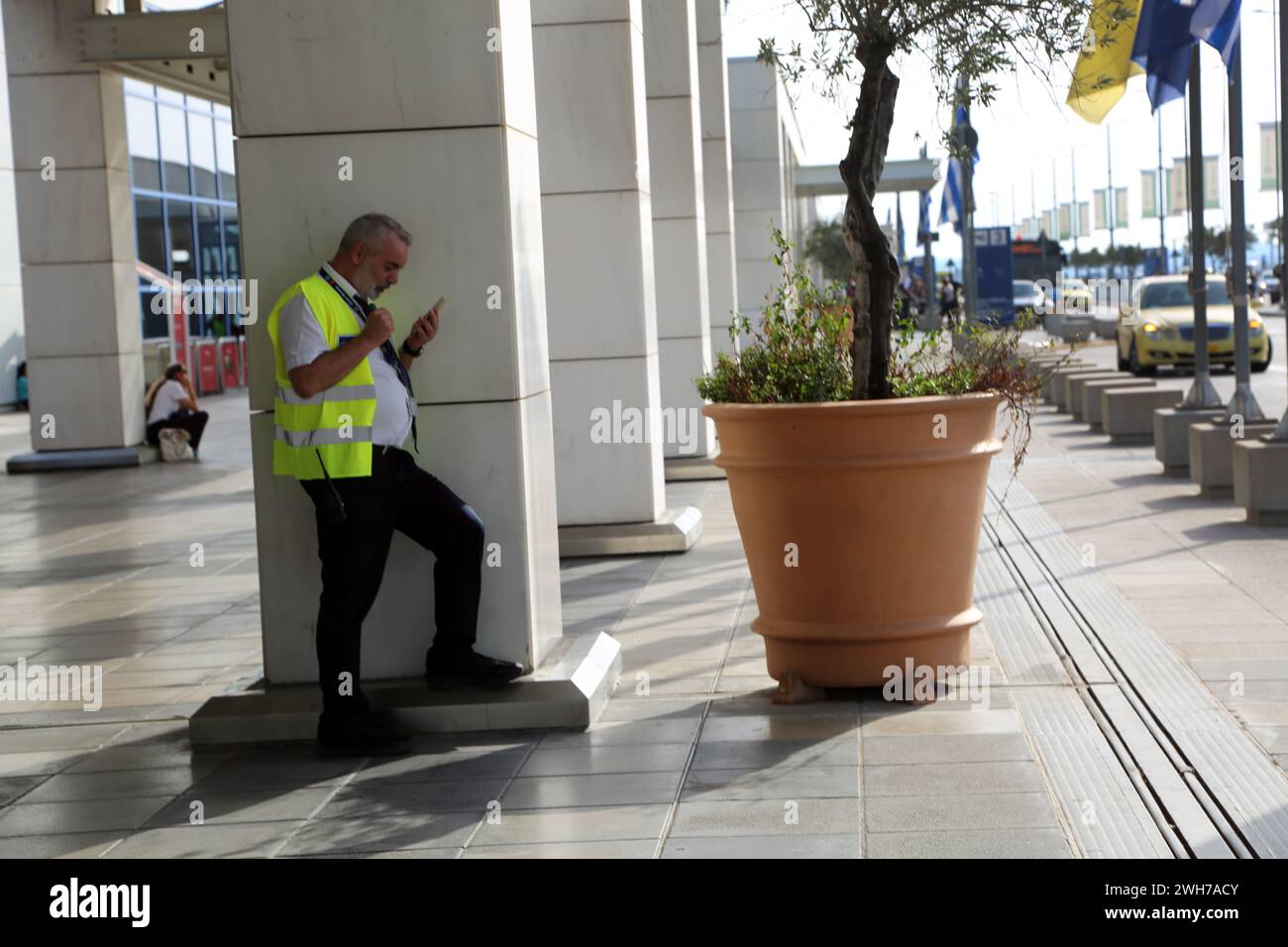 Atene Grecia Aeroporto Internazionale di Atene (AIA) Eleftherios Venizelos Man Wearing High Visibility Gilet Standing Olive Tree in Pot Outside Foto Stock