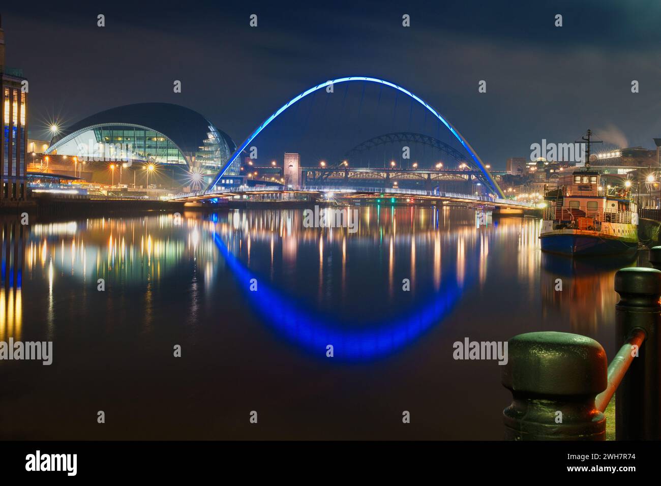 La banchina di Newcastle e i ponti Tyne di notte con riflessi di luce nel fiume Tyne. Una barca è ormeggiata al molo e la Glass House è sulla. Foto Stock