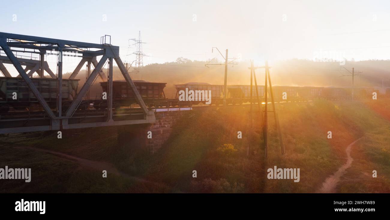 Il treno merci attraversa il ponte ferroviario attraverso il fiume all'alba. Il sole del mattino basso illumina le carrozze. Foto Stock