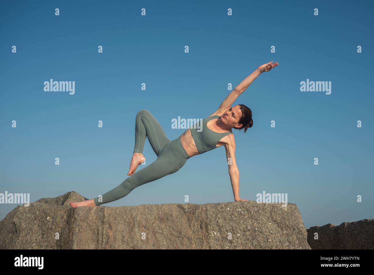 donna in forma che fa una posa di yoga su rocce con tavola laterale, sfondo blu del cielo Foto Stock