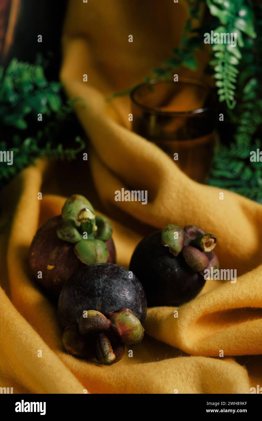 Spettacolare natura morta con tre mangostani viola, viti verdi, un vivace panno giallo in feltro e un bicchiere d'acqua. Primo piano della dolce fru tropicale Foto Stock