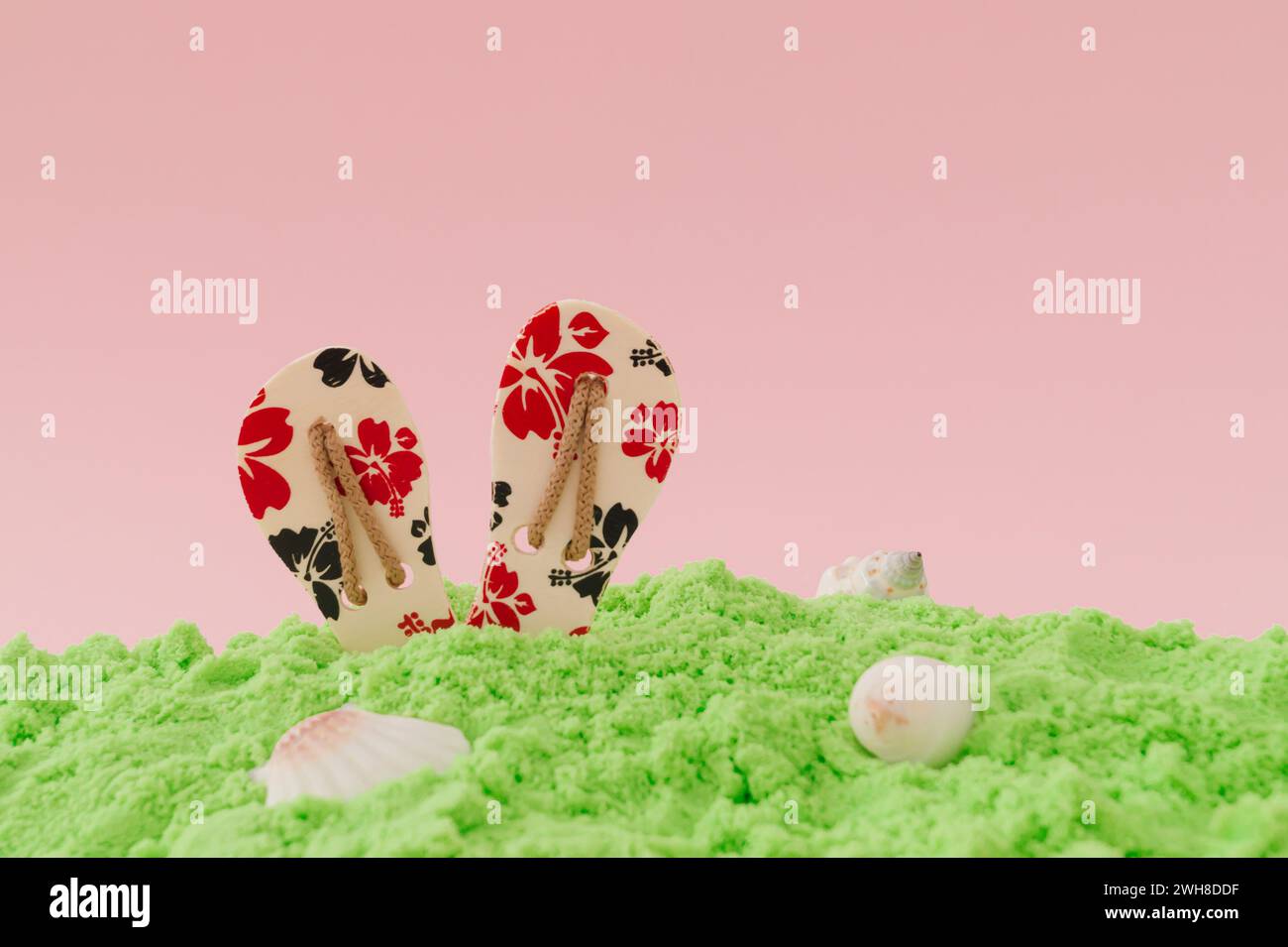 Elegante e magica disposizione estiva realizzata con pantofole floreali, sabbia verde da spiaggia e conchiglie marine su sfondo rosa chiaro. Concetto creativo minimalista. Foto Stock