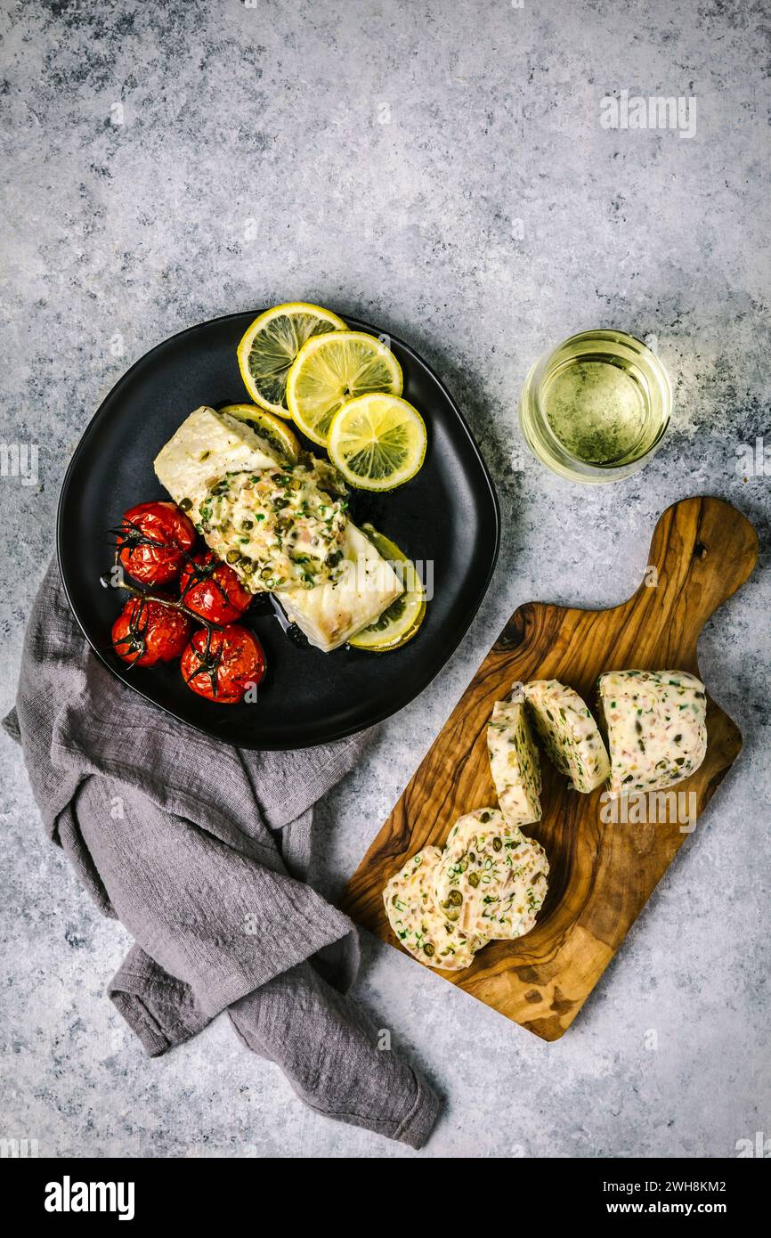 Colpo di testa con filetto di pesce bianco ricoperto di burro composto fuso su un piatto nero con pomodori arrostiti, limoni a fette e vino bianco in un gambo Foto Stock