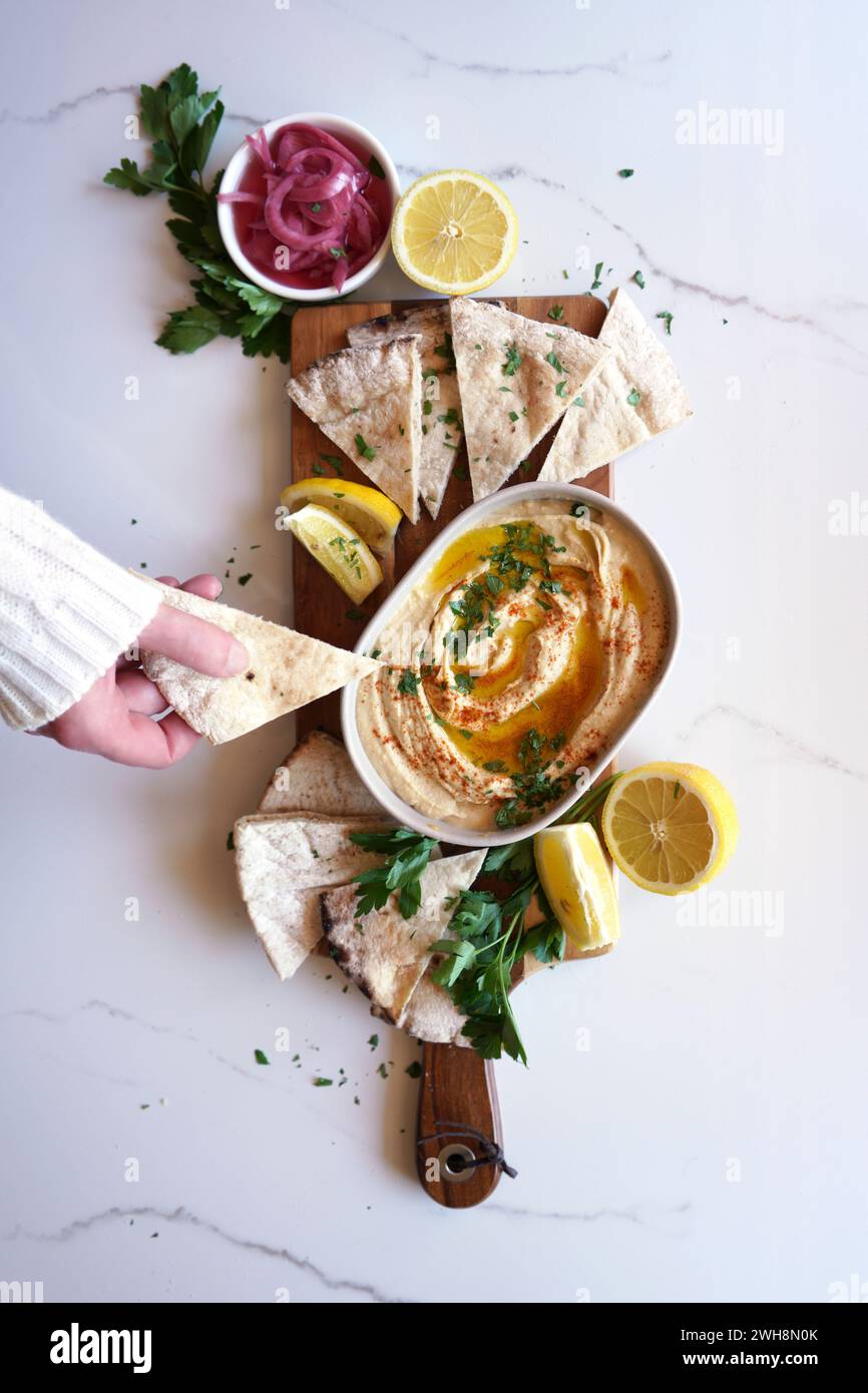Hummus di ceci servito con olio d'oliva, cipolla rossa sottaceto, limoni e focaccia su un tavolo di marmo bianco. Focaccia che si immerge a mano femminile. Foto Stock